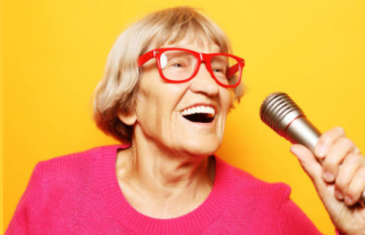 Older woman wearing a pink jumper and red glasses happily singing into a microphone 