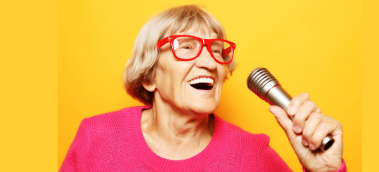 Older woman wearing a pink jumper and red glasses happily singing into a microphone 