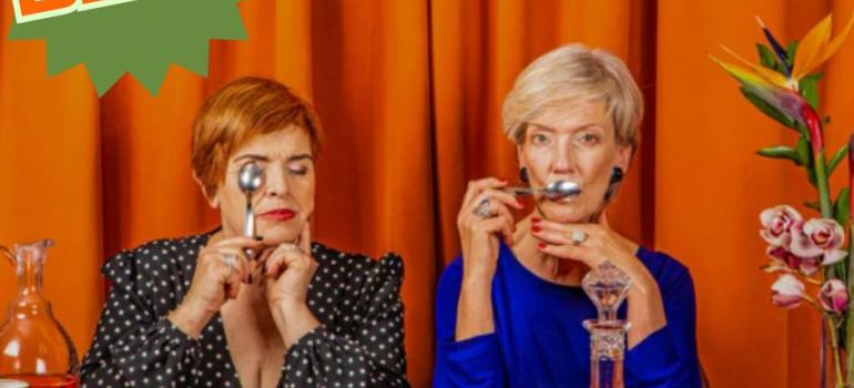 2 women sitting at a table with cake and wine in front of an orange curtain